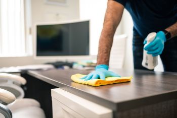 Person wiping a desk with spray cleaner in an office workspace